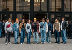 Skupina mladých lidí před vchodem do koncertní haly v neformálním oblečení, s rušným pozadím.