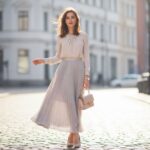 Stylová žena v pastelové plisované sukni a blůze stojí na dlážděné ulici v městském prostředí.
