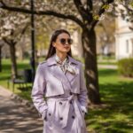 Mladá žena v lehkém trenčkotu a šátku kráčí městským parkem s kvetoucími sakurami.