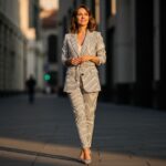 Žena v pruhovaném saka a úzkých kalhotách stojí na městské ulici za teplého denního světla.
