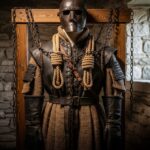 Historický katovský kostým na dřevěném stojanu v místnosti s kamennými zdmi a řetězy.