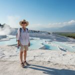 Turista na terasách Pamukkale v Turecku v létě s kloboukem a láhví vody, termální bazény v pozadí.